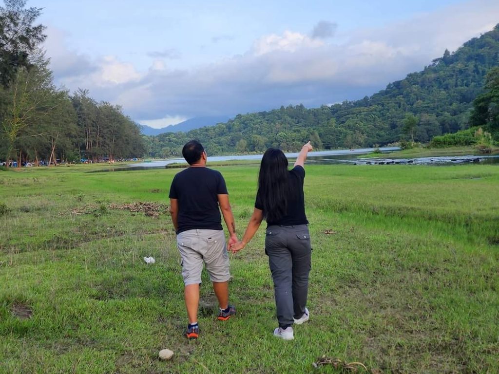 Pair in casual wear holding hands and looking at a scenic lake and hills.