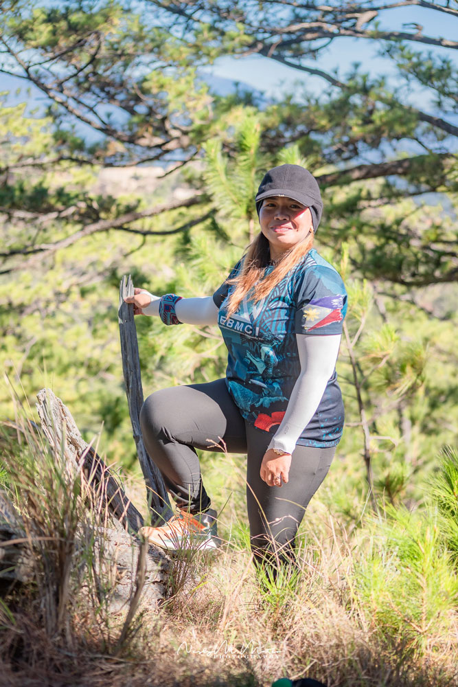 Smiling hiker in gear rests on a fence amidst greenery.